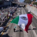 A vibrant demonstration featuring a large Mexican flag being waved by a protester, with a crowd of people holding signs gathered along a freeway underpass.